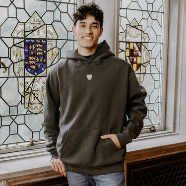 College-aged man wearing the Sigma Alpha Epsilon embroidered black hoodie, standing in front of stained glass windows with fraternity-style crest designs.