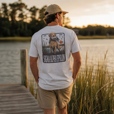 Young man wearing a white graphic T-shirt while standing on a wooden dock overlooking a lake at sunset.