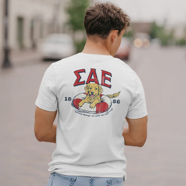 Sigma Alpha Epsilon  fraternity member in a limited edition  white short sleeve tee  featuring a golden retriever puppy playing in an pool floatie  with the fraternity name written above and the founding year.