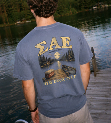 Young man wearing a blue graphic tee standing on a lakeside dock, showing the back design