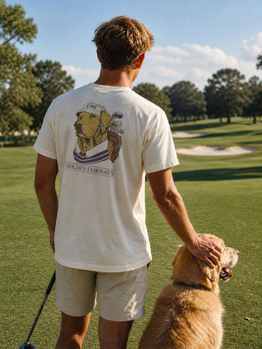 Lifestyle photo of a college student wearing a golf-themed fraternity t-shirt on a golf course with a golden retriever. Ivory Comfort Colors tee features a “Golden Fairways” back graphic with a golden retriever in golf apparel and golf clubs, perfect for fraternity golf tournaments, campus events, and casual Greek life apparel.
