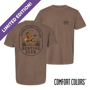 Brown garment-dyed short-sleeve tee featuring a vintage-inspired graphic: a weathered oval frames a mallard in mid-flight with burnt orange and mossy green-tipped wings above rippling water and cattails; distressed Sigma Alpha Epsilon arcs overhead and SAE letters bold and rugged on the front.
